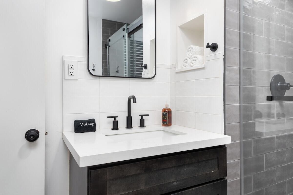 Un baño con un lavabo blanco, grifo negro y un espejo encima. Hay un neceser sobre el lavabo y una botella de jabón en la encimera.