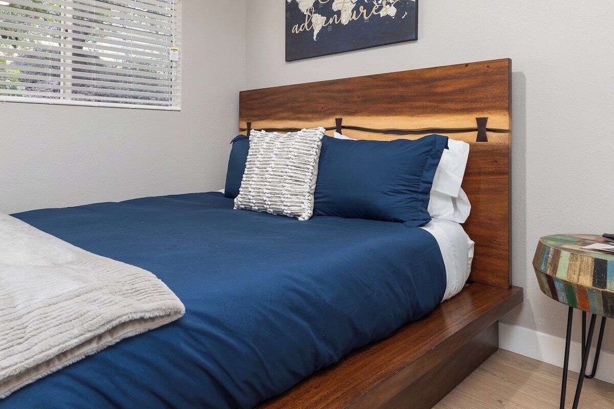 A neatly made bed with a wooden headboard, a world map on the wall, and a small round table with a colorful top.