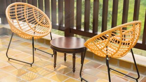 Dos sillas de mimbre con estructura metálica y una pequeña mesa redonda en un patio empedrado, con una barandilla de madera al fondo.