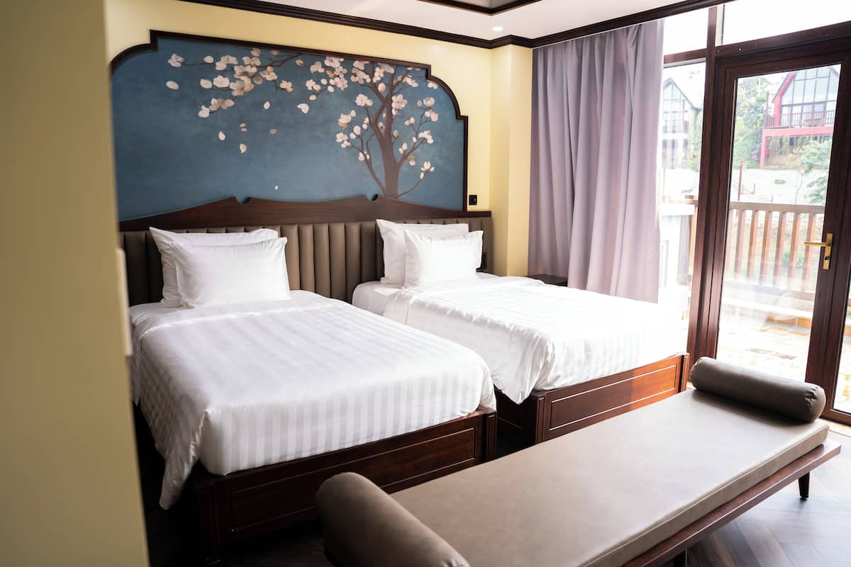 Habitación de hotel con dos camas, un banco y un ventanal con cortinas.
