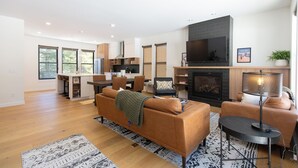 A modern living room with a sofa, a coffee table, and a fireplace. There is a dining area with a table and chairs, and a kitchen in the background. - Alpine Nest Bridger Bowl A (Bozeman)