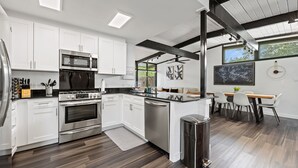 A modern kitchen with white cabinets, stainless steel appliances, and a dining area with a wooden table and chairs. - Stylish Home near Love-Field Airport, King & Queen Beds with a Large Yard (Dallas)