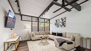 A modern living room with a sectional sofa, a glass coffee table, a floor lamp, and a large flat-screen TV mounted on the wall. - Stylish Home near Love-Field Airport, King & Queen Beds with a Large Yard (Dallas)