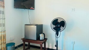 A room with a flat-screen TV mounted on the wall, a small table with a refrigerator and a kettle, a standing fan, and a trash bin. - Ella Nature Valley Homestay- Family Friendly Lovely Place surround by mountains. (Ella)