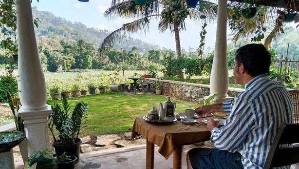 A man sitting at a table with a view of a lush garden and hills in the background. - Ella Nature Valley Homestay- Family Friendly Lovely Place surround by mountains. (Ella)