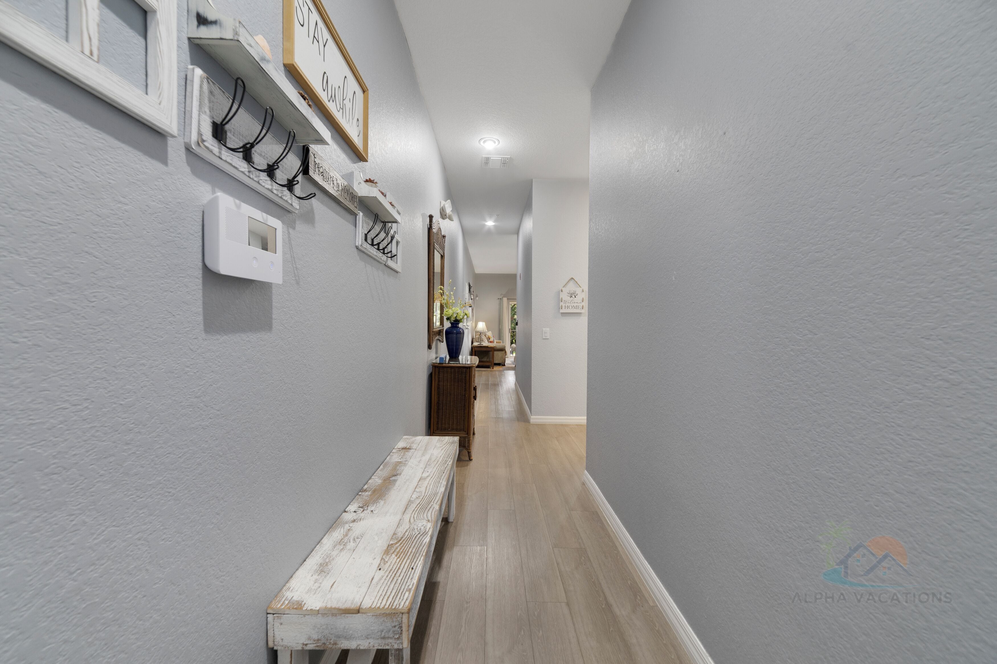 Un couloir avec un banc, des patères fixées au mur, un thermostat et un panneau encadré.
