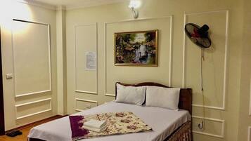 Un dormitorio ordenado con una cama, un cuadro en la pared y un ventilador de pie.