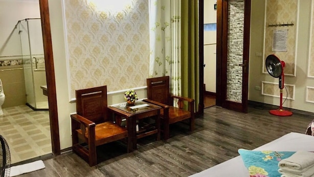 Une chambre d’hôtel avec un plancher en bois, un lit recouvert d’une courtepointe à motifs floraux, une petite table avec des chaises, un ventilateur sur pied et une salle de bain visible à travers une porte ouverte.