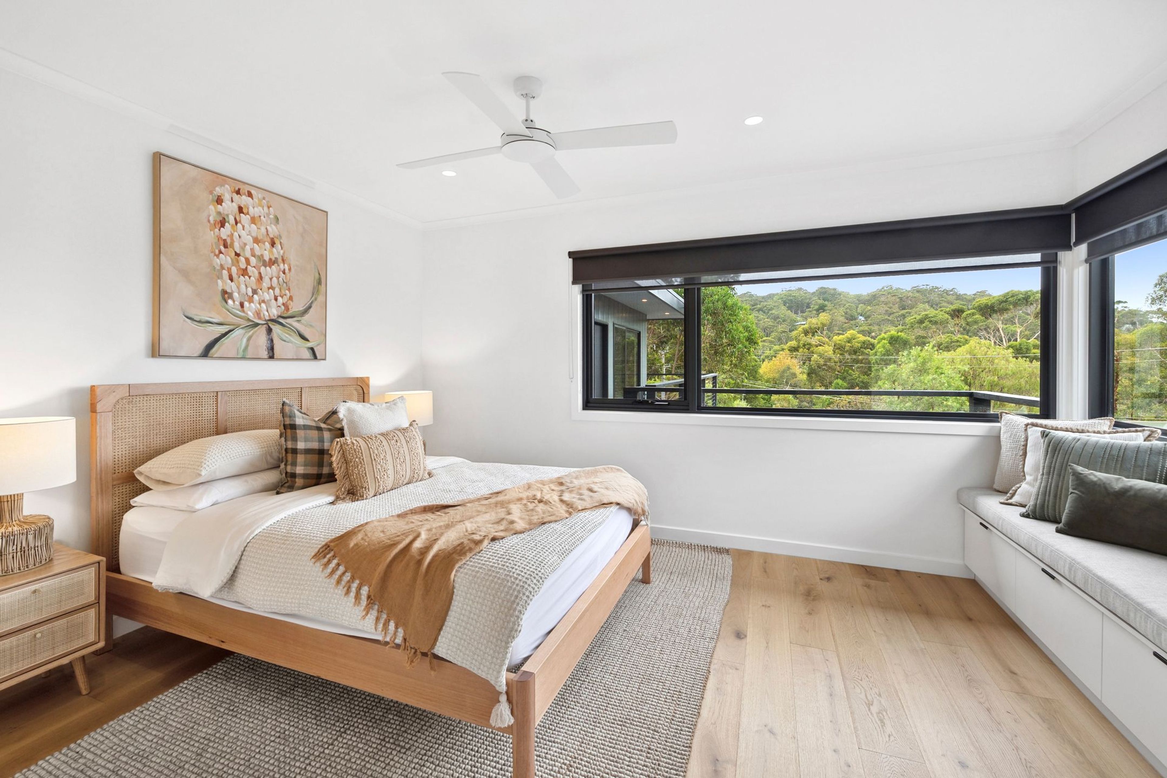 A bedroom with a large bed, a ceiling fan, a window with a view of greenery, and a bedside table with a lamp.