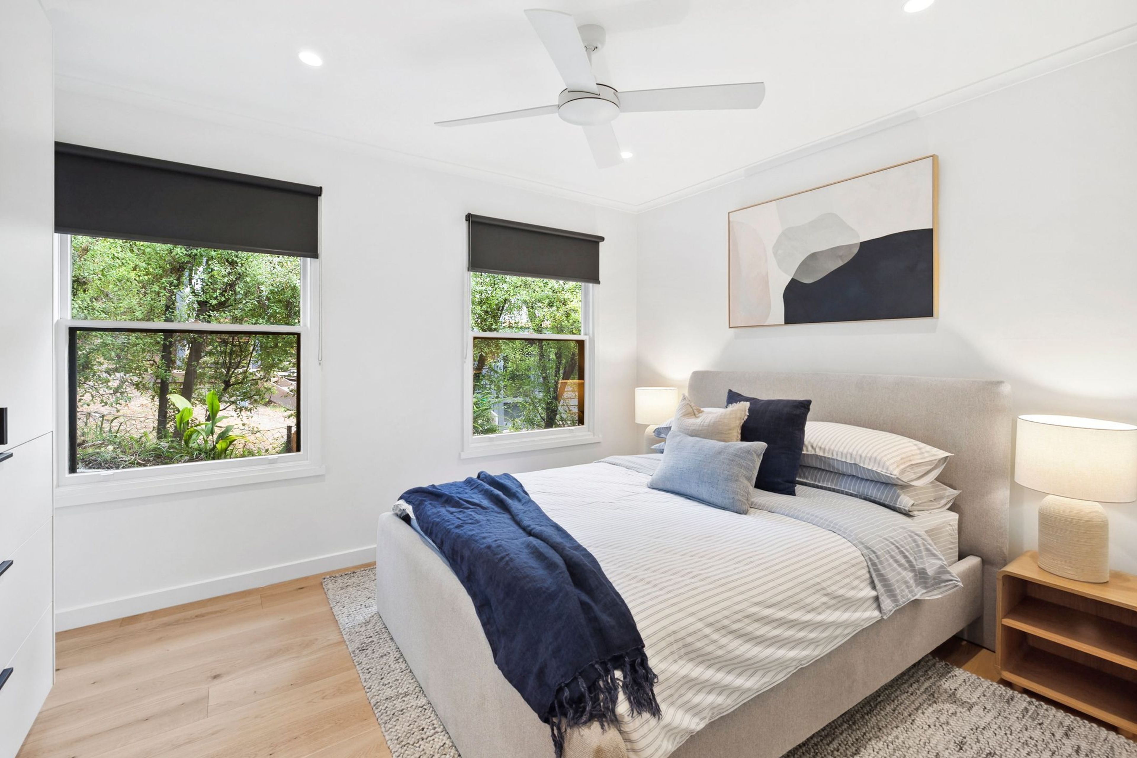 A bedroom with a bed, two bedside lamps, a ceiling fan, and two windows with blinds.