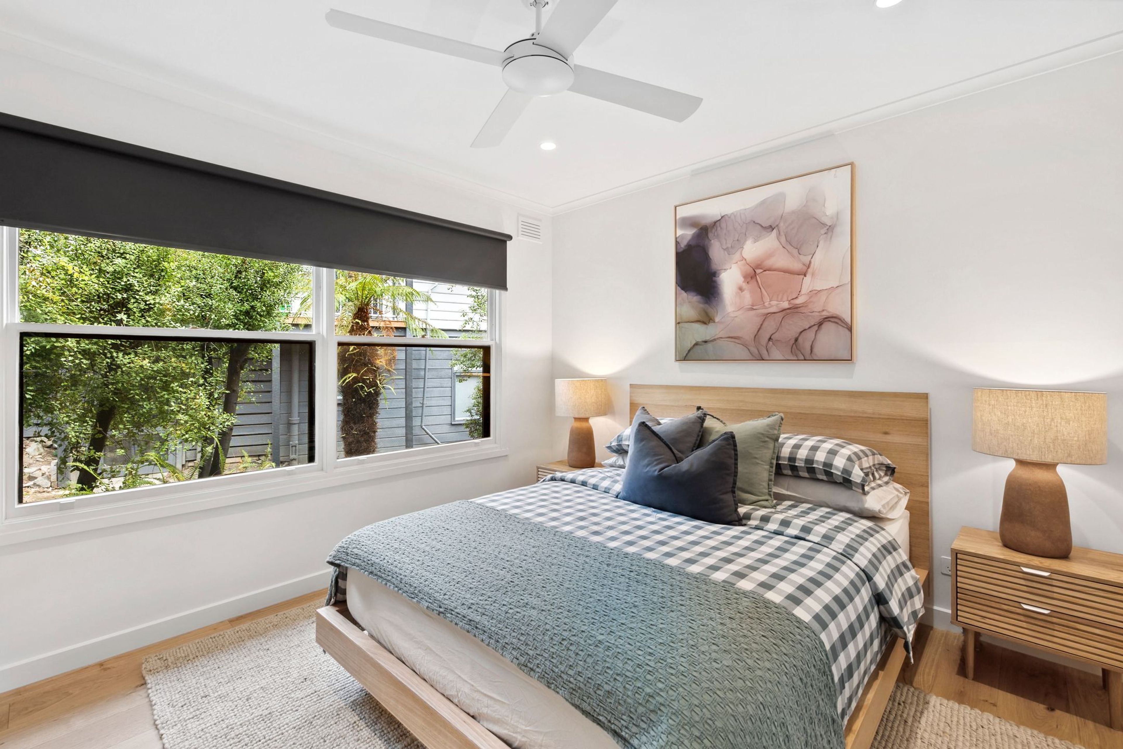 A bedroom with a bed, two bedside lamps, a ceiling fan, a window with a view of greenery, and a wall-mounted abstract painting.