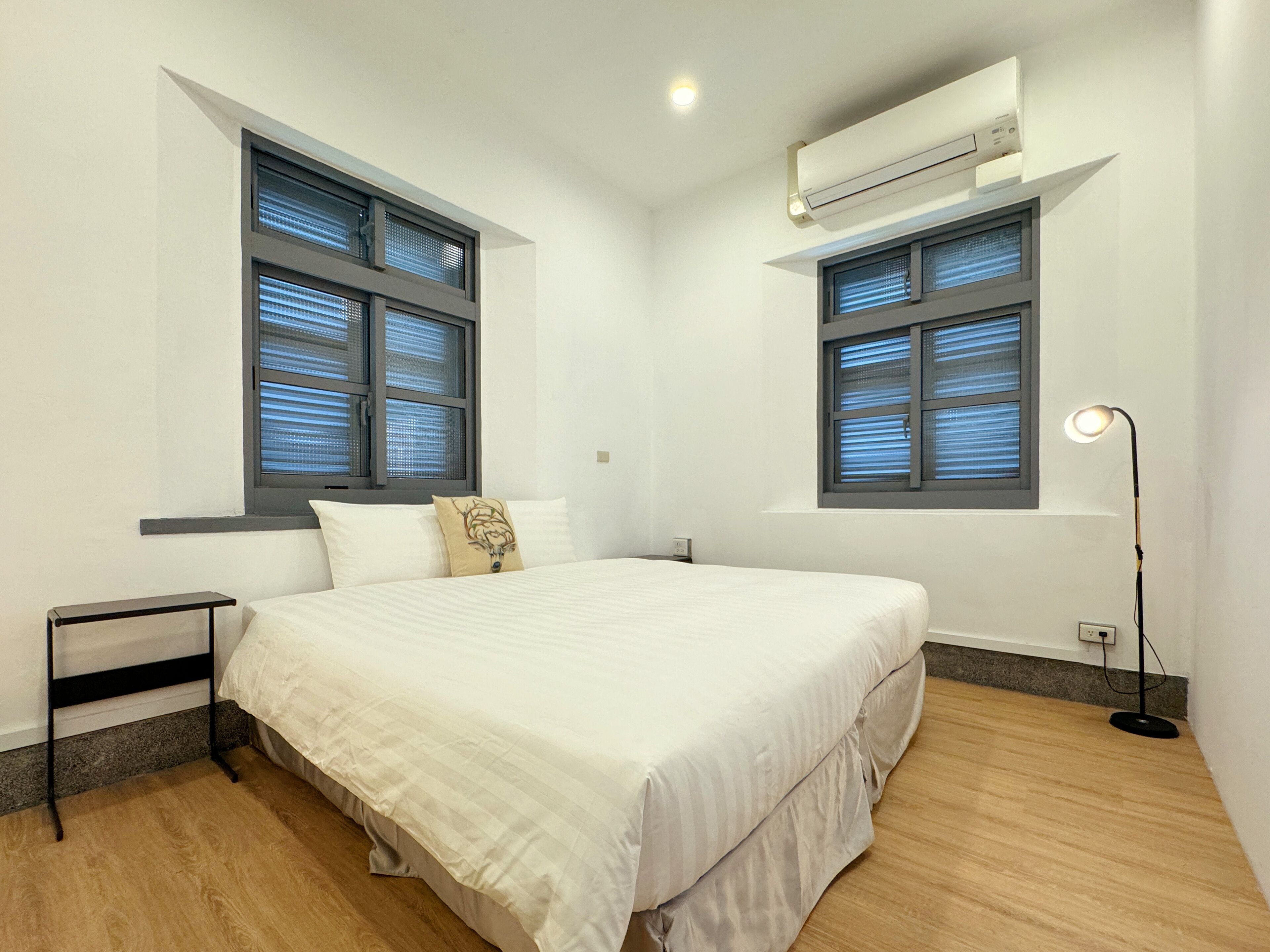 Un dormitorio con cama, dos ventanas con persianas, una mesita de noche, una lámpara y un equipo de aire acondicionado.