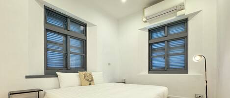 A bedroom with a bed, two windows with blinds, a bedside table, a lamp, and an air conditioning unit.