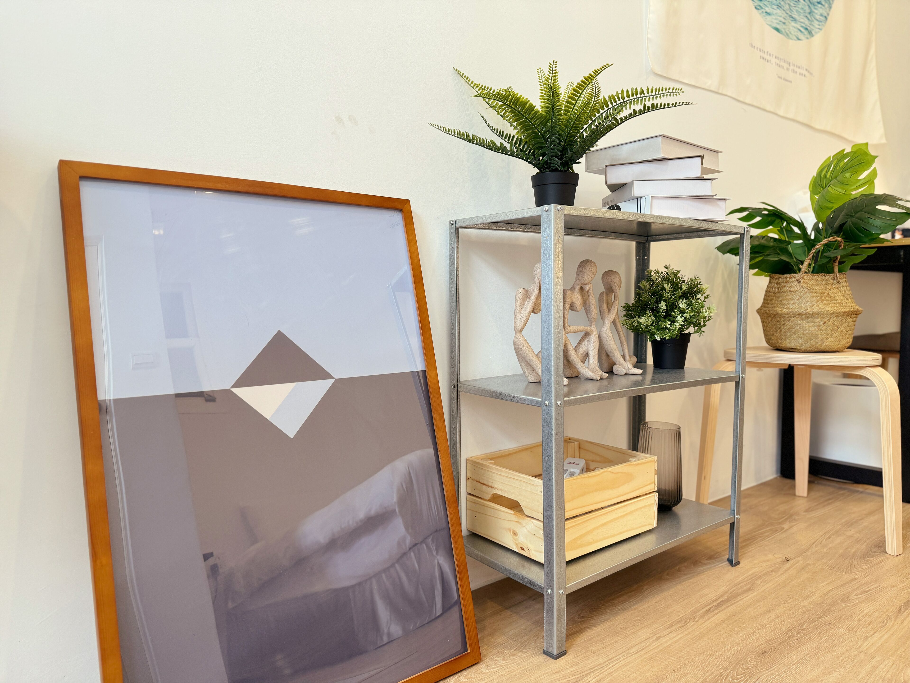 Un cuadro enmarco, una estantería de metal con libros y plantas, y una silla de madera.