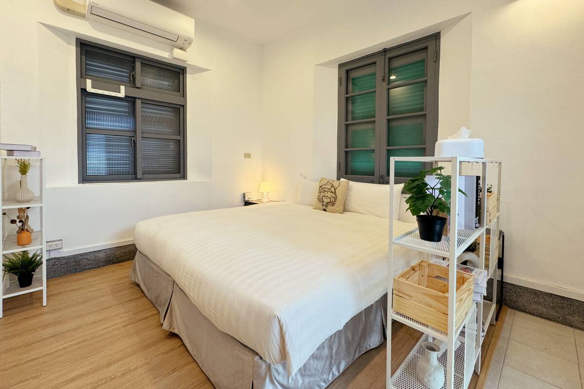 A neatly arranged bedroom with a large bed, a bedside table with a lamp, a window with blinds, a shelf with plants, and a white wardrobe.