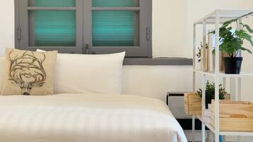 A bedroom with a bed, a window with green blinds, a white shelf with plants, and a decorative pillow.