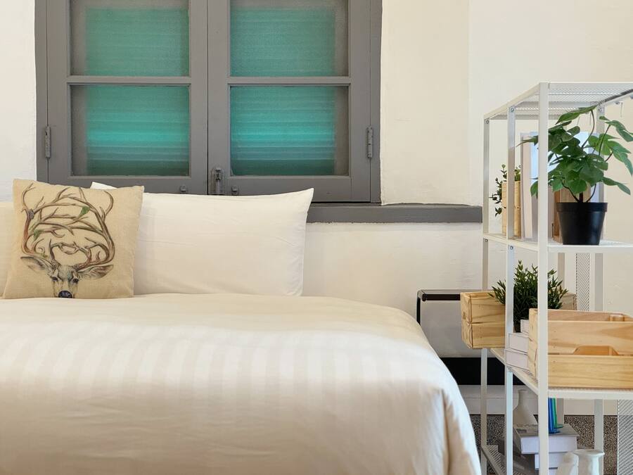 Un dormitorio con una cama, una ventana con persianas verdes, una estantería blanca con plantas y un cojín decorativo.