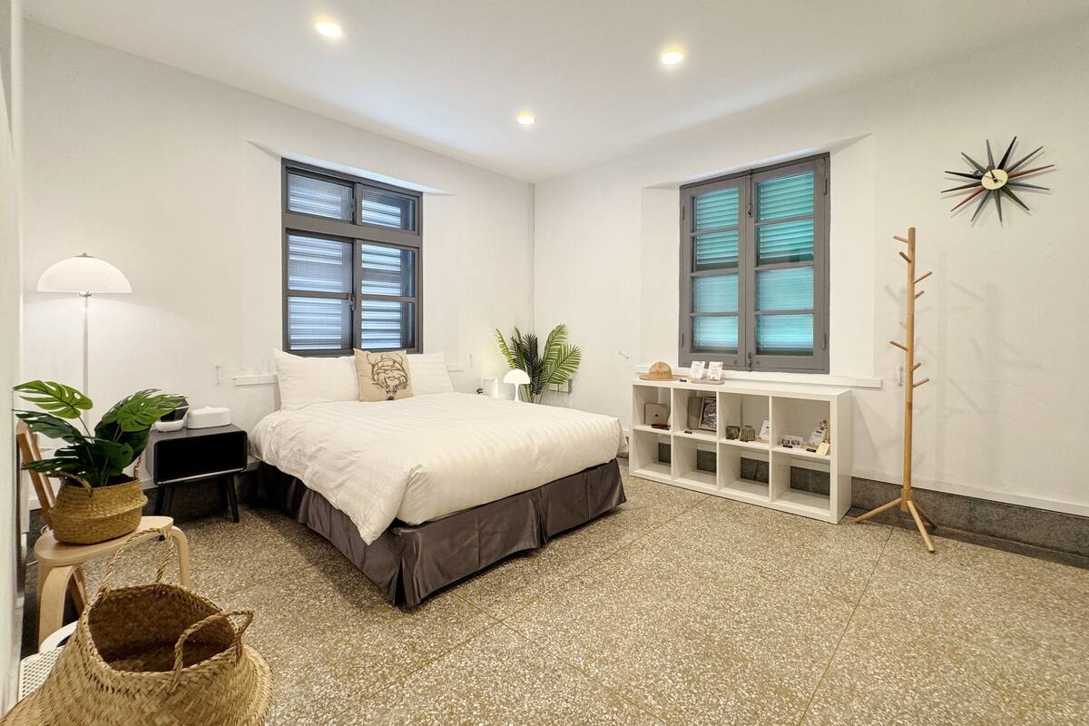 A bedroom with a bed, two windows, a white console table, a floor lamp, and a plant.
