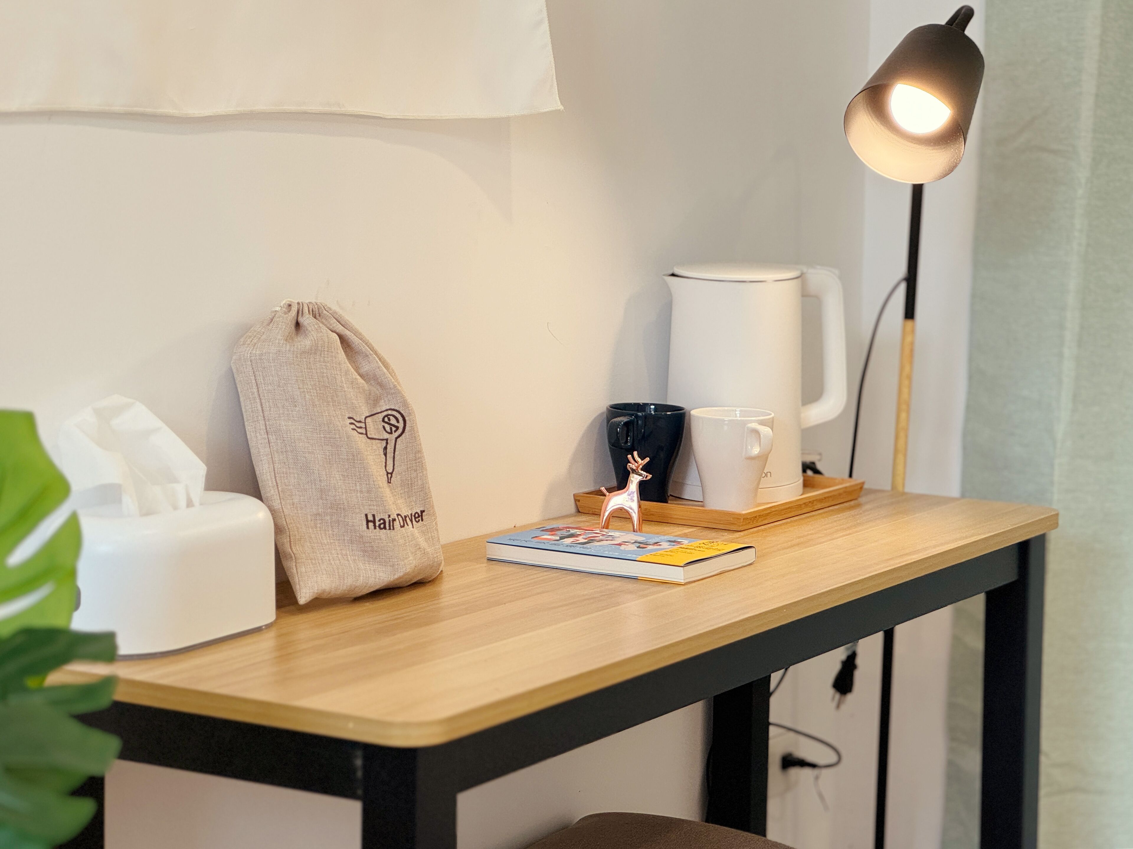 A small wooden table with a coffee maker, a mug, a book, a hairdryer bag, and a lamp.