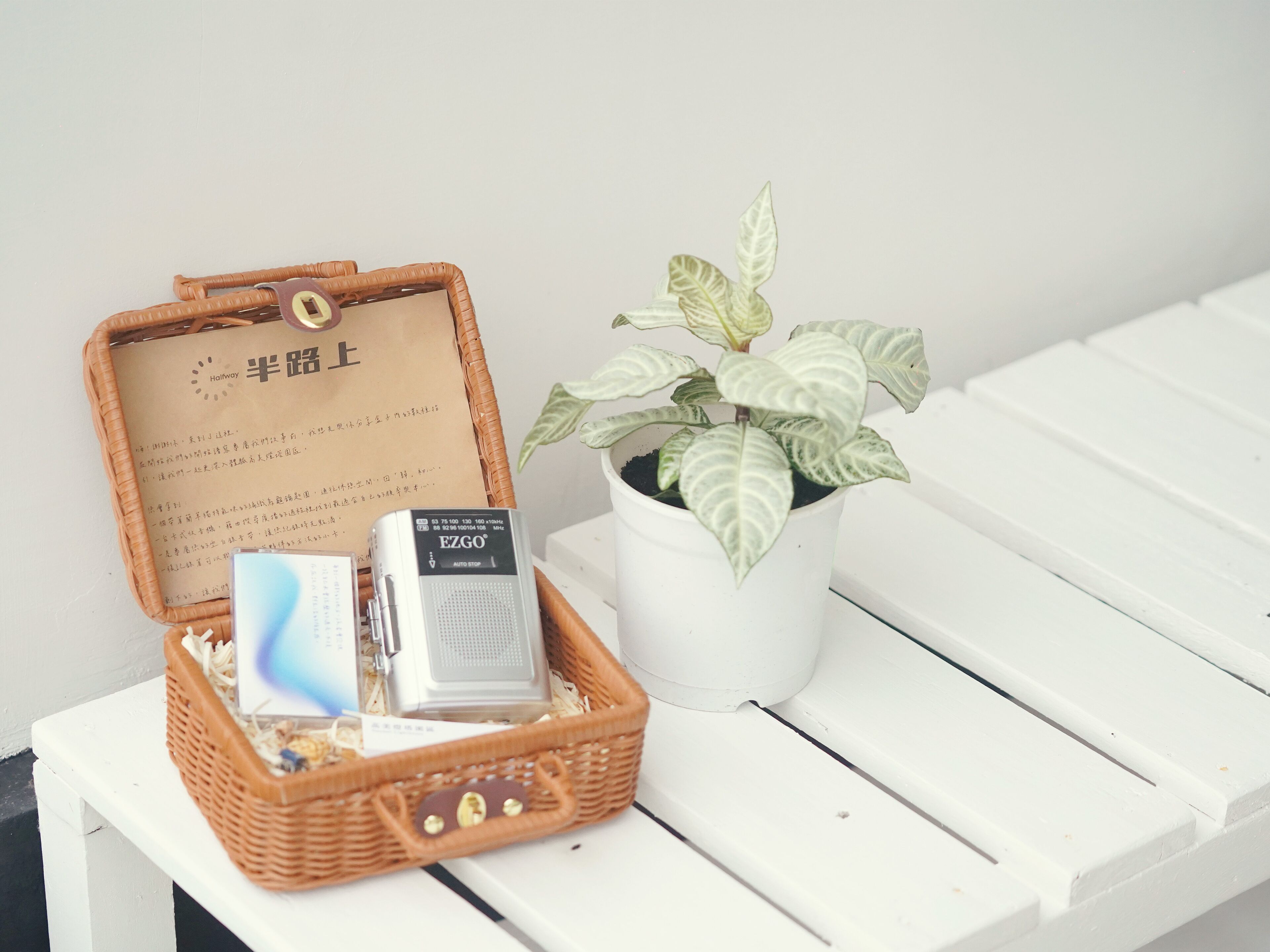 A wicker basket with a card and a device, next to a potted plant on a white bench.