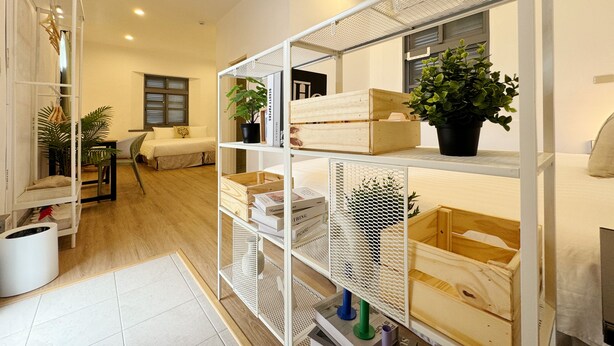 Una habitación moderna y bien iluminada con una estantería metálica blanca que contiene diversos objetos, una cama con cabecera blanca y una ventana con persianas.