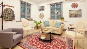 Una sala de estar con un sofá, dos sillones, una mesa de centro y un tapete. Hay ventanas, un reloj y un perchero con una chaqueta.