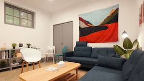 Una sala moderna con sofá, mesa de centro de madera y una silla blanca. Se observa una estantería con libros y objetos decorativos, un cuadro abstracto de gran tamaño en la pared y una puerta al fondo.