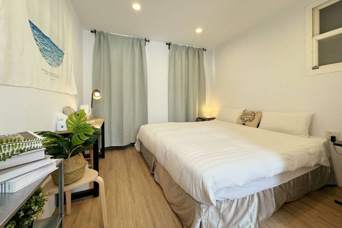 A hotel room with a bed, a desk with a lamp, a chair, and a plant.