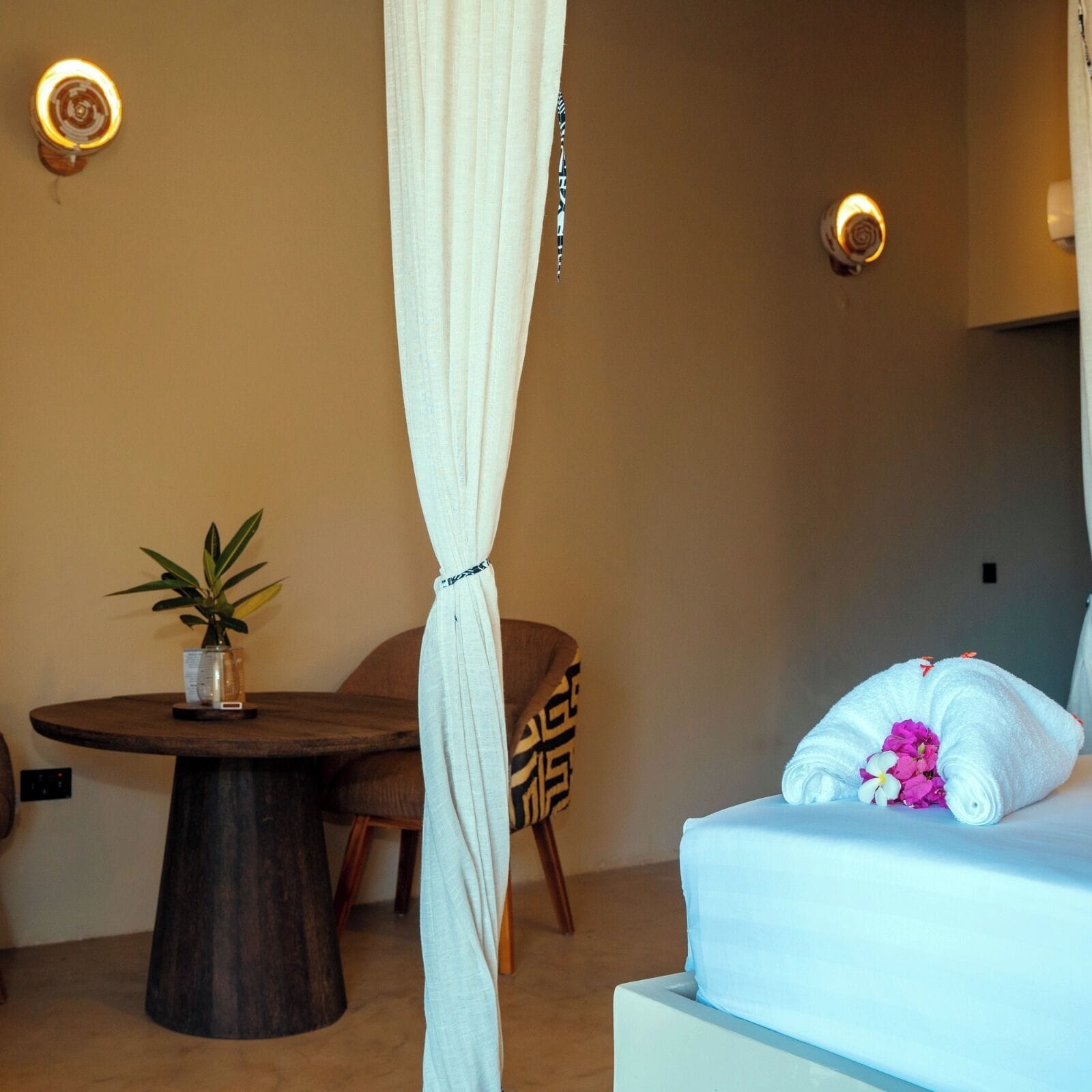 A room with a bed, a small round table with a plant, and a chair. There are two wall-mounted lights and a curtain.