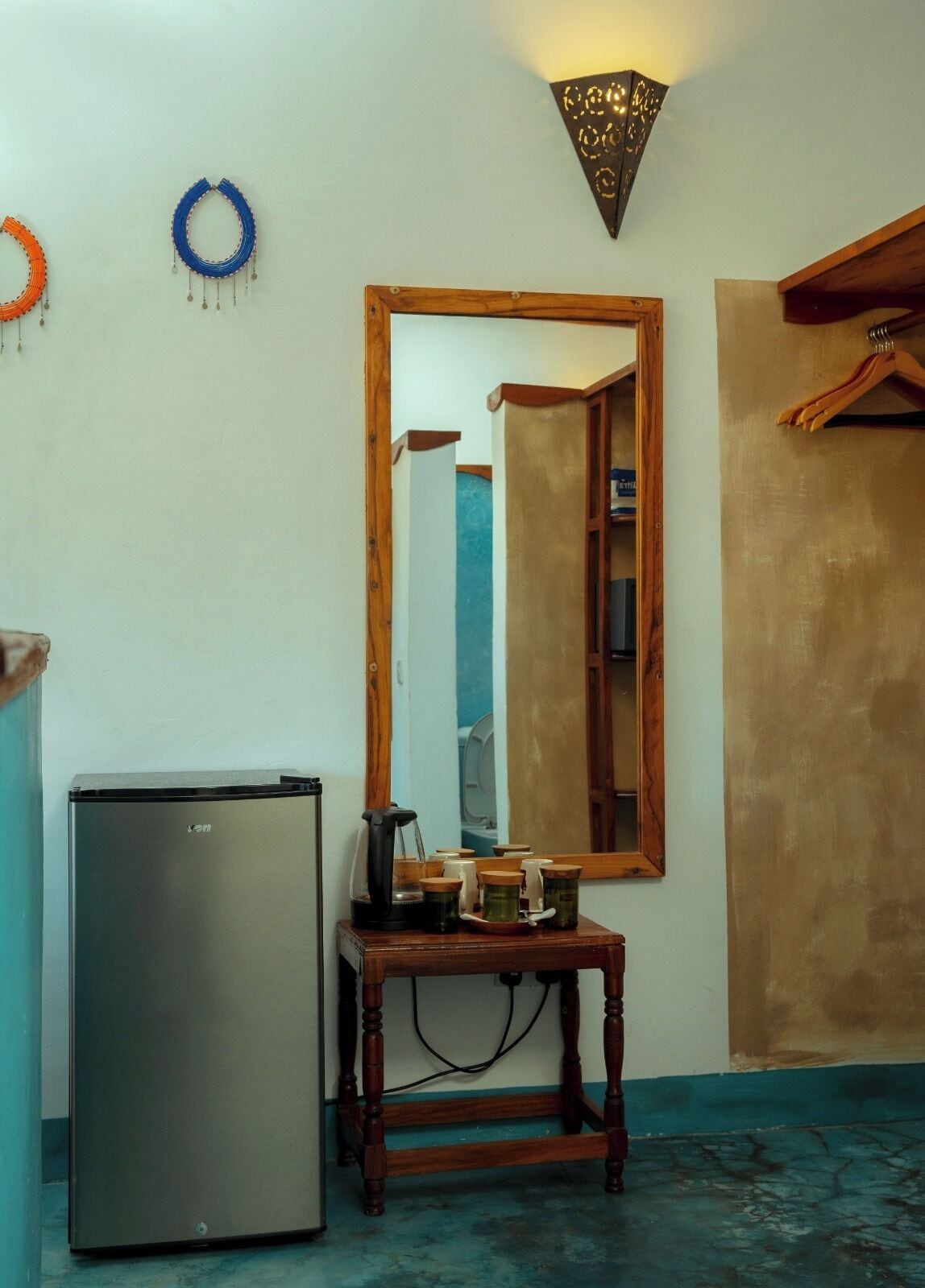 A compact room with a refrigerator, a wooden table with jars, a mirror, and a wall-mounted light fixture.