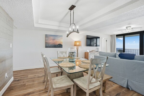 A dining area with a glass table, wooden chairs, a chandelier, and a view of the ocean through large windows.