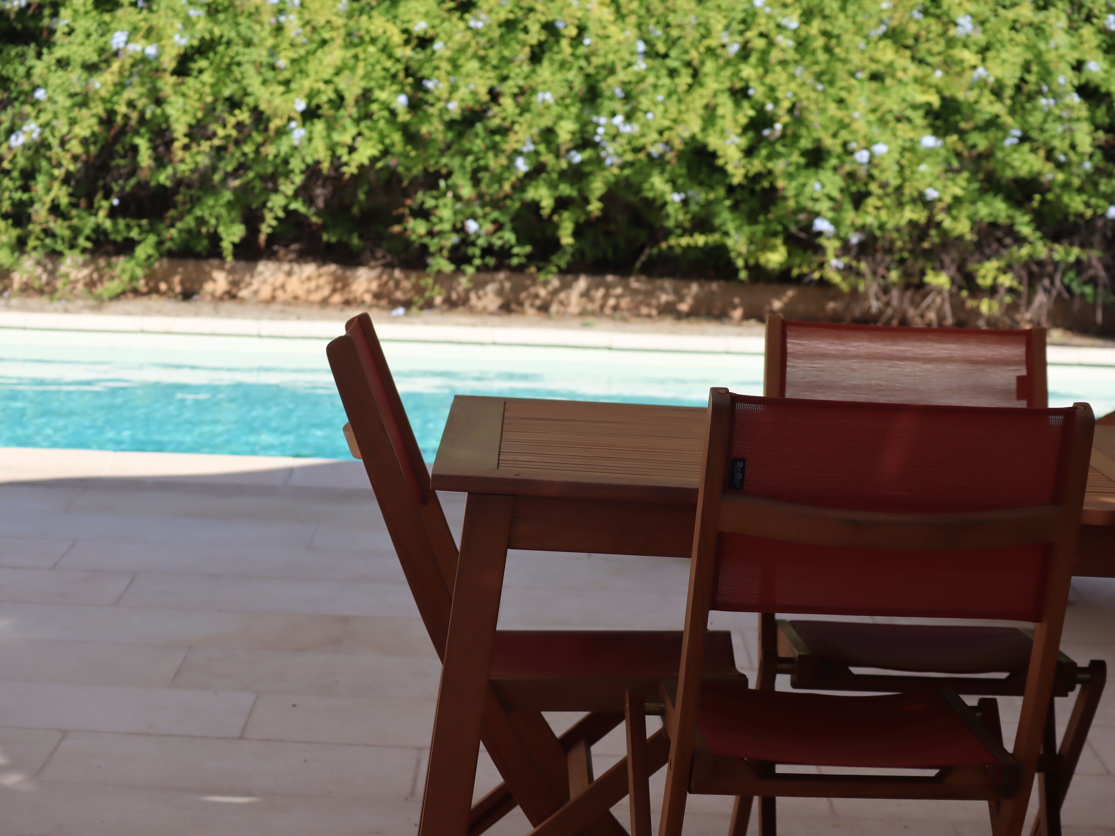 A wooden table and chairs by a poolside with a view of a lush green hedge.