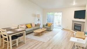 A modern living room with a sofa, dining table, and chairs. There is a fireplace, a TV mounted on the wall, and a sliding glass door leading to an outdoor area. - Family-Friendly Retreat in Marysville (Marysville)