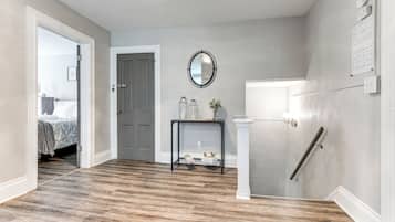 Un couloir doté d’un plancher en bois, d’une porte blanche, d’une petite console ornée d’objets décoratifs, d’un miroir fixé au mur et d’une main courante sur le côté droit.