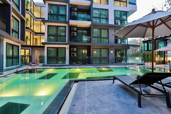 A modern hotel with a swimming pool, lounge chairs, and a well-lit exterior.