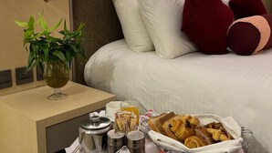 A hotel room with a neatly made bed, a bedside table with a tray of breakfast items, a potted plant, and a wall-mounted lamp. - FALAJ DARIS HOTEL (Nizwa)