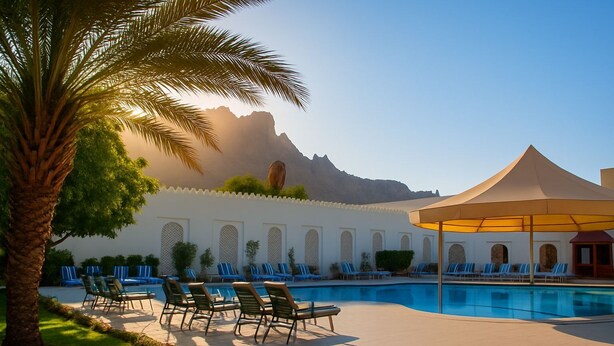 A pool area with lounge chairs and a tent, surrounded by a white wall with arches and a mountain in the background. - FALAJ DARIS HOTEL (Nizwa)
