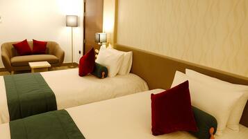 A hotel room with two beds, each with white linens and red pillows. There is a sofa with a red cushion and a small table beside it. A floor lamp is visible in the corner.
