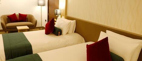 Habitación de hotel con dos camas, cada una con sábanas blancas y almohadas rojas. Hay un sofá con un cojín rojo y una mesita auxiliar a su lado. Se ve una lámpara de pie en la esquina.
