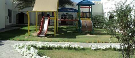 Un parque infantil con tobogán y estructuras de escalada, rodeado por un muro blanco y palmeras.