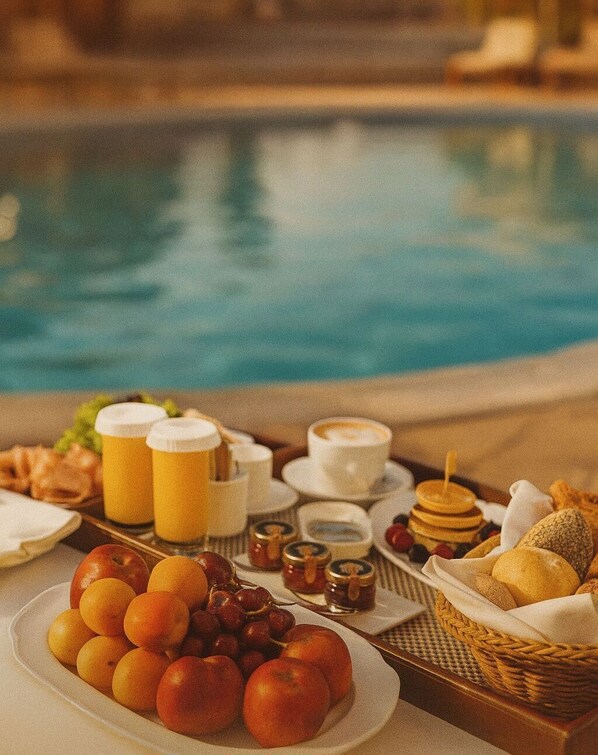 A breakfast setup by a pool with fruits, pastries, and beverages.