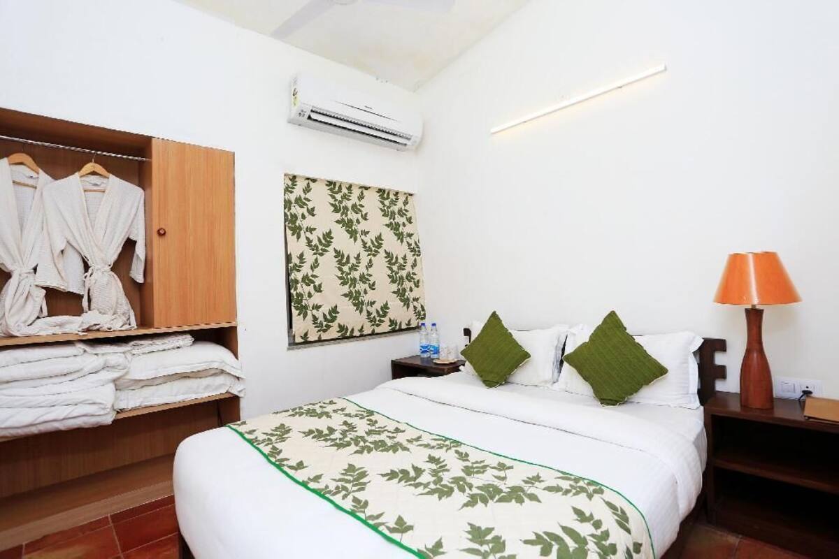 A hotel room with a bed, a wardrobe, a wall-mounted air conditioner, a bedside table with a lamp, and a green leaf-patterned wall hanging.