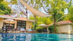 A swimming pool with lounge chairs and umbrellas, surrounded by trees and a building with large windows. - Aranya Nivas Resort (Samakhunta)