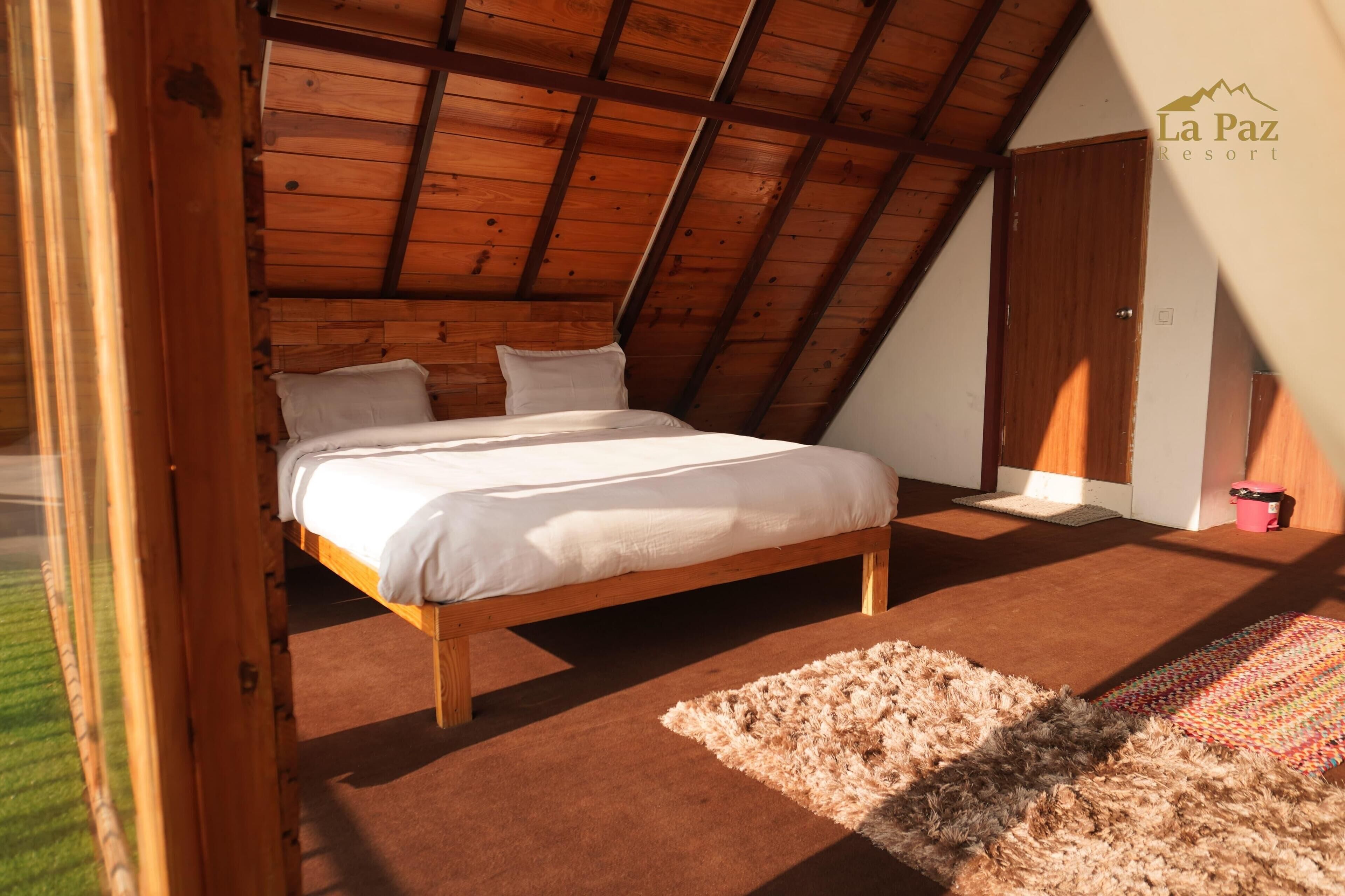 A bedroom with a wooden ceiling, a bed with white bedding, a fluffy rug, and a door to the right.