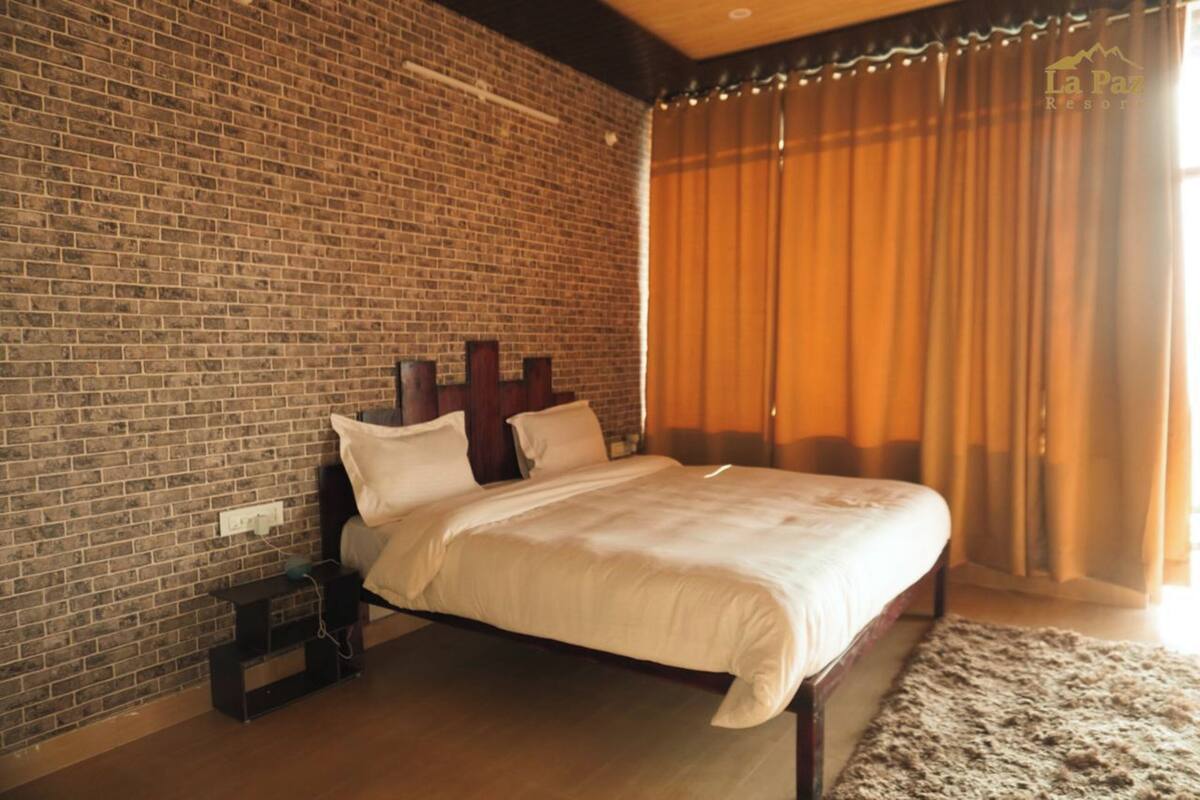 Une chambre avec un mur en briques, une tête de lit en bois, un lit avec une literie blanche et une grande fenêtre avec des rideaux.