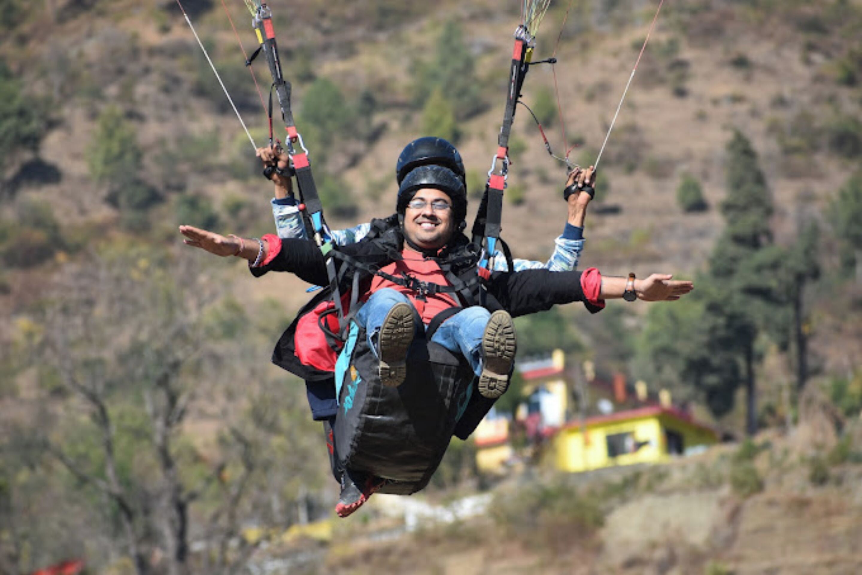 A person paragliding with a harness and safety gear, set against a backdrop of a hilly landscape and a distant yellow building.