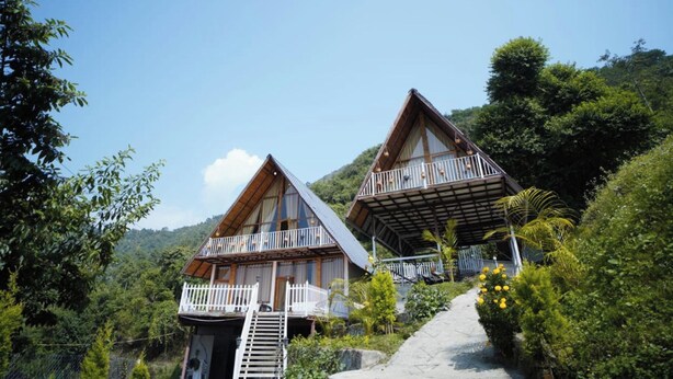 Two wooden cabins with balconies, surrounded by lush greenery and a clear sky. - La Paz Resort By Shikhar Hospitality (Nainital)