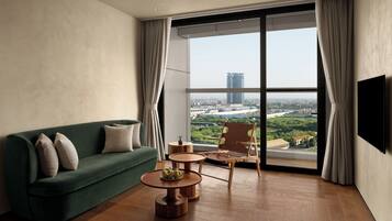 Una sala de estar moderna con un sofá verde, una mesa de centro de madera y una silla con vistas a un paisaje urbano a través de ventanas amplias.