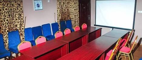 A conference room with a long table, blue and pink chairs, patterned curtains, a projector screen, and a ceiling fan.
