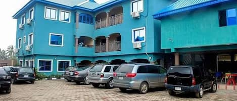 A multi-story building with a blue exterior, a parking area with several cars, and a balcony on the upper floor.